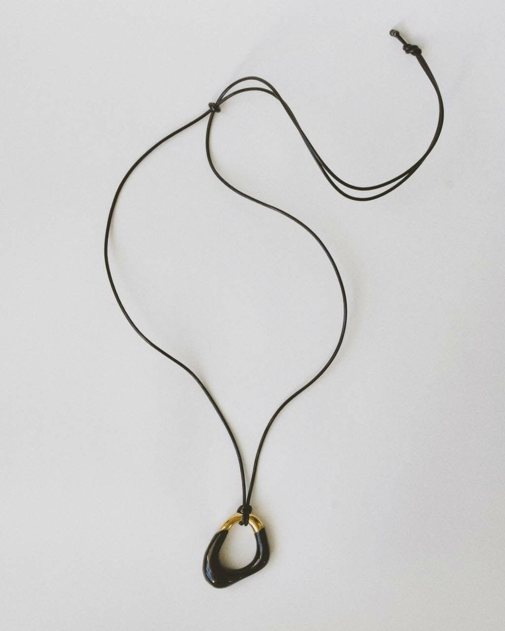 Momentum Necklace in black and gold by Enso Design Lab, displayed flat with a vegan leather cord. The pendant's unique, liquid-inspired design is highlighted with enamel and gold plating.