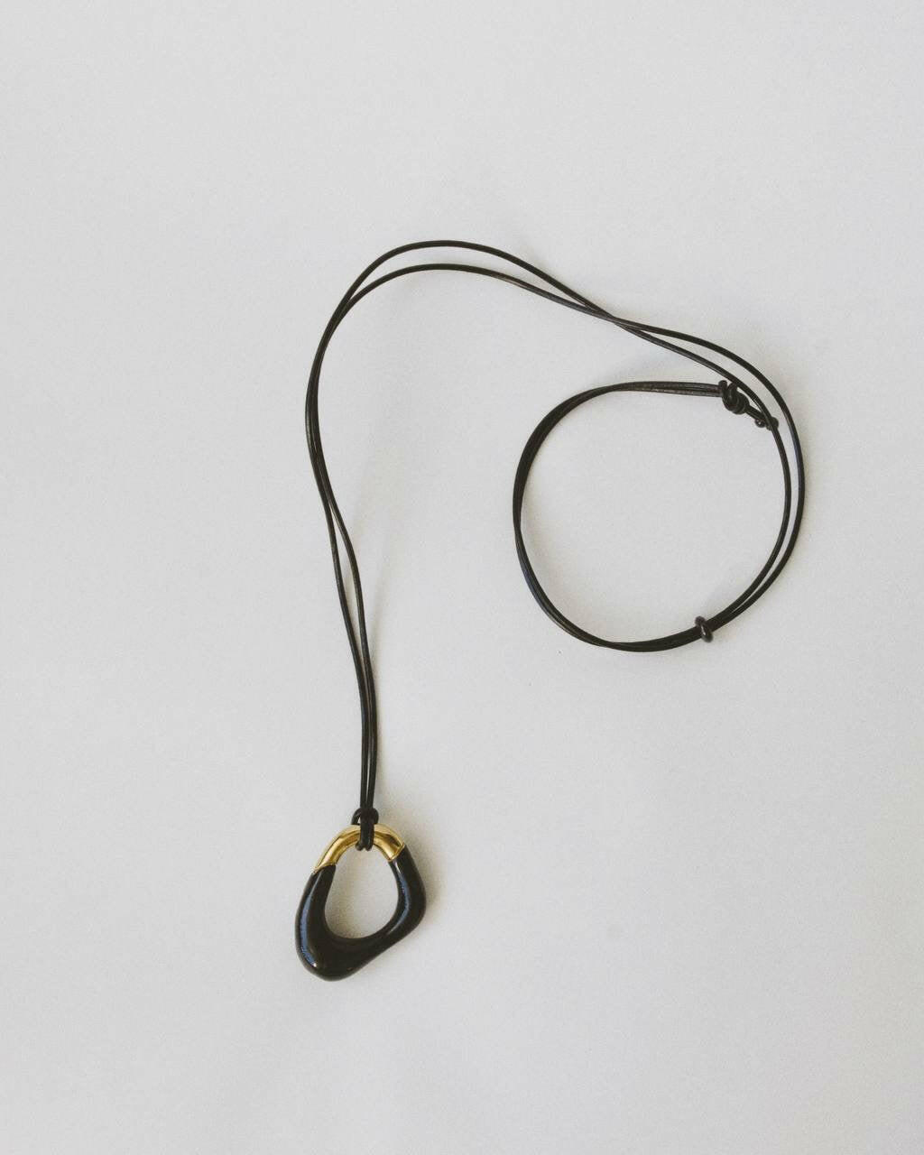Close-up of the Momentum Necklace in black and gold, showing the pendant and vegan leather cord. The design features smooth, flowing lines inspired by surrealism, with enamel and gold plating.