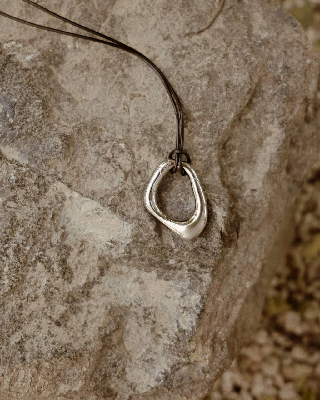 Close-up of the Momentum Necklace by Enso Design Lab, designed by Ani Han, resting on a stone surface. The pendant's silver finish and fluid form are highlighted.