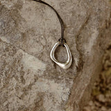Close-up of the Momentum Necklace by Enso Design Lab, designed by Ani Han, resting on a stone surface. The pendant's silver finish and fluid form are highlighted.