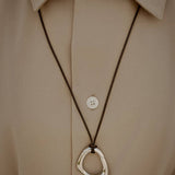 Close-up of the Momentum Necklace in silver color, designed by Ani Han, worn over a beige shirt. The pendant hangs on a leather cord, showcasing its liquid-inspired design.