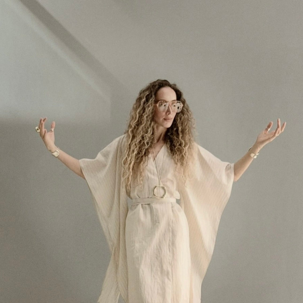 Ani Han, creative director of Enso Design Lab, standing in a light kaftan with open arms against a soft grey wall.