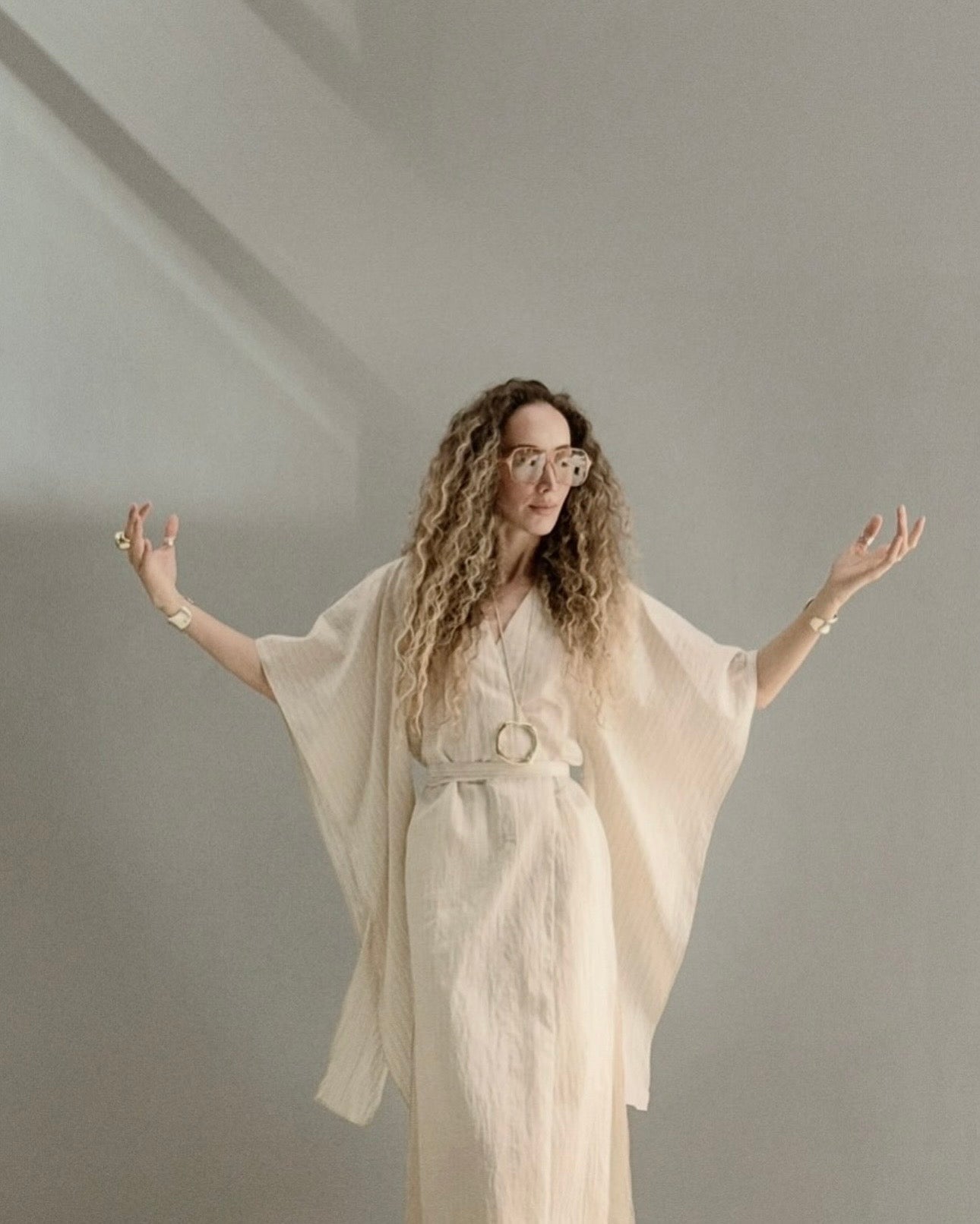 Ani Han, creative director of Enso Design Lab, standing in a light kaftan with open arms against a soft grey wall.