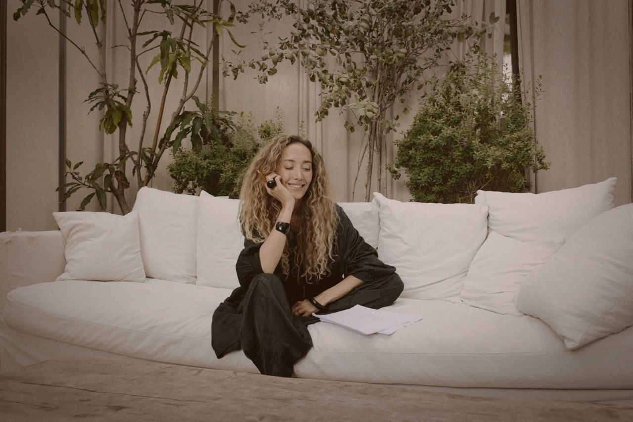 Ani Han, the creative director of Enso, sitting on a white sofa surrounded by greenery, smiling while talking on the phone and holding papers.