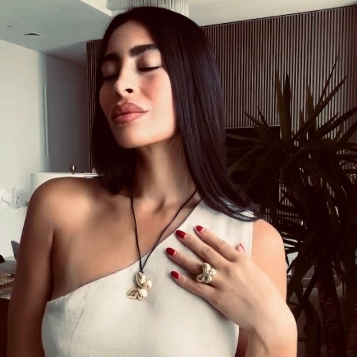 Nour Ahmed (@nour.moussad) gently touches her chest showcasing a delicate gold necklace and matching ring from Enso Design Lab’s Metamorphosis collection. Her expression is serene, highlighting the luxurious and thoughtful design of the jewelry.