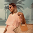 A woman, Marie Engie, happily wearing sunglasses and a striped pink and white dress, holding a woven bag, standing in a sunny outdoor setting with palm trees in the background. She embodies the lifestyle promoted by Enso Design Lab, reflecting joy and satisfaction with their sustainable products.