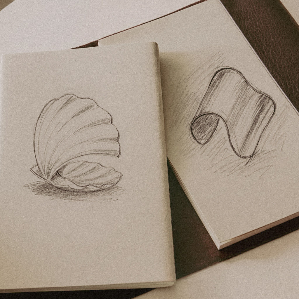Two open sketchbooks with pencil drawings of a shell and abstract shapes on a wooden surface. Technical sketch and Shell Cuff bracelet showing design development